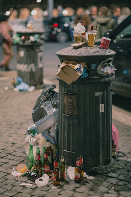 A row of five large, white plastic waste bins with closed lids positioned on a paved outdoor area. To the left, there are two smaller black bins labeled 'PAPERS' and 'CARDBOARD ONLY,' each with closed