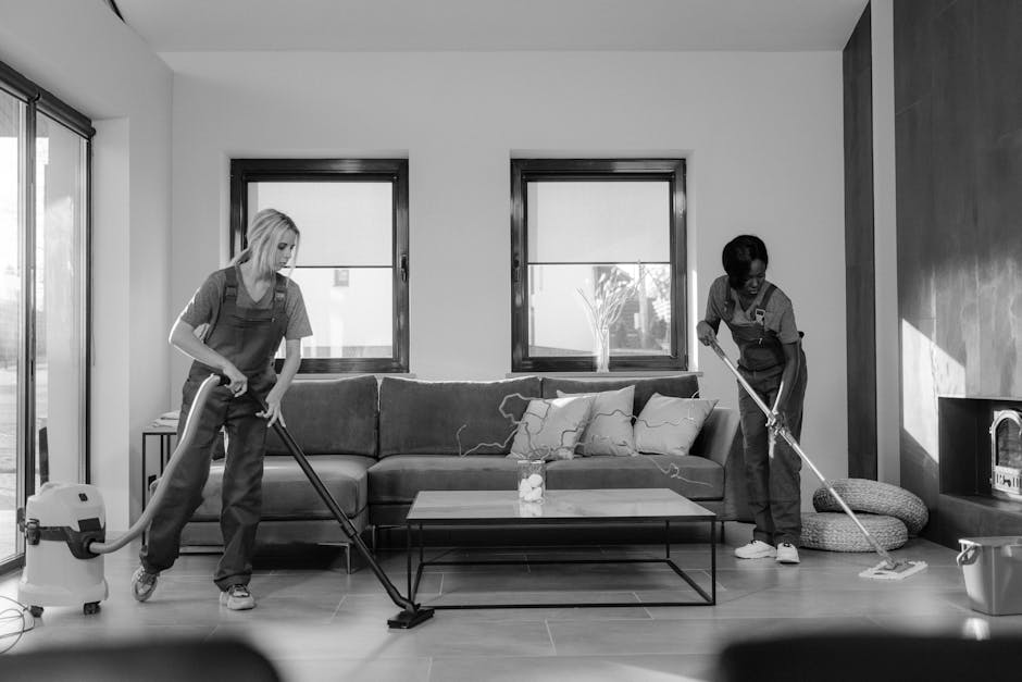 Two professional cleaners from Deep Cleaning Ealing standing back-to-back in a well-lit, clean indoor space with a plain, light-colored wall background. The cleaner on the left, with dark hair tied ba
